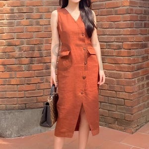 May include: A sleeveless, rust-colored dress with a V-neck and button-down front. The dress features two front pockets and a slit at the hem. The person is wearing sunglasses and holding a handbag.