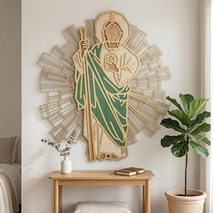 May include: Wooden wall art depicting a religious figure with a halo, holding a staff and a medallion. The figure is dressed in white and green robes, set against a gold sunburst design. The artwork is mounted on a white wall above a wooden table.