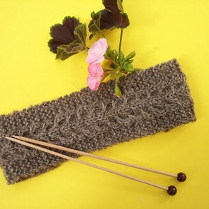 May include: A brown knitted headband with a cable knit pattern, lying on a yellow surface with knitting needles and pink flowers.