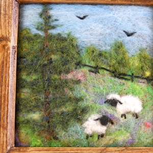 May include: A needle felted landscape art piece in a brown wooden frame featuring two white sheep with black faces grazing in a green meadow. The scene includes a tall evergreen tree, a black fence, and two black birds flying in a blue sky.