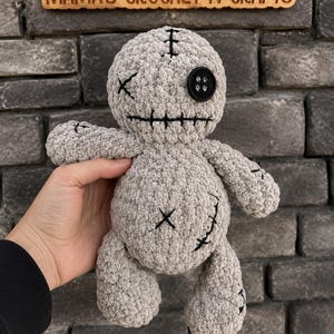 Könnte beinhalten: Eine graue Plüschpuppe aus strukturiertem Garn mit rundem Kopf und Körper. Das Gesicht hat ein schwarzes Knopfauge, ein gesticktes X-förmiges Auge und einen gestickten horizontalen Mund. Der Körper hat ein gesticktes X und eine gebogene Linie. Text MAMA'S CROCHET N CRAFTS.