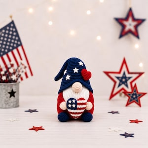 May include: A crochet gnome in red, white, and blue, holding a heart-shaped design with the American flag. The gnome wears a blue hat with white stars and a red heart. Decorative stars and an American flag are also present.