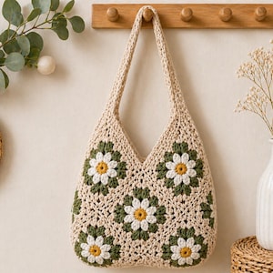 May include: A cream-colored crochet tote bag with daisy flower patterns in green, white, and yellow. The bag is hanging on a wooden peg rack. A vase with dried flowers and a woven coaster are also visible.