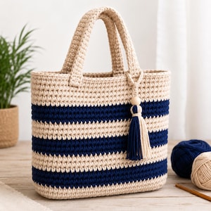 Könnte beinhalten: Eine gehäkelte Tragetasche mit beigen und marineblauen Streifen und passenden Griffen. Die Tasche hat eine dekorative Quaste mit einer Holzperle. Die Tasche steht auf einer Holzoberfläche.