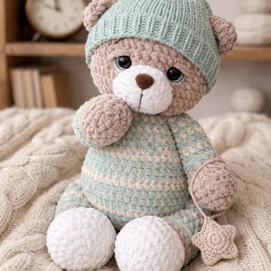 Könnte beinhalten: Ein handgehäkelter Teddybär mit hellblauer Mütze und weißem Bommel. Der Bär hat einen hellbraunen Körper, blaue Augen und eine weiße Schnauze. Er hält ein sternförmiges Spielzeug und sitzt auf einer cremefarbenen Decke.