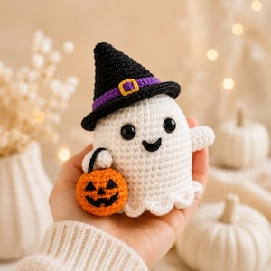 May include: A white crocheted ghost figurine wearing a black witch hat with a purple band and a yellow buckle. The ghost has black safety eyes, a stitched smile, and holds a small orange crocheted pumpkin bucket with a black jack-o'-lantern face.