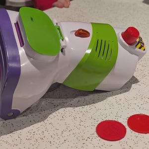 Könnte beinhalten: Ein weiß-grün-violetter Spielzeug-Blaster aus Kunststoff mit einem roten Knopf auf der Oberseite, einer grünen Klappe und einem kleinen orangefarbenen Licht. Zwei separate rote, runde Scheiben mit Sternprägung liegen vor dem Spielzeug.