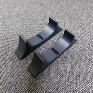 May include: Two black, curved stands with a rectangular cutout in the center. The stands have a smooth finish and are designed to hold an item. The stands are made of a solid material and are placed on a gray surface.