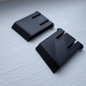 May include: Two black, 3D-printed objects with a rectangular base and a notched top. The objects are stacked, showing a layered texture. The items are on a white surface, with the top object angled towards the viewer.