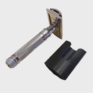 Puede incluir: Una maquinilla de afeitar de seguridad cromada con un mango texturizado y una funda protectora negra. La maquinilla tiene un diseño clásico, con una cabeza rectangular y un agarre moleteado detallado para un manejo seguro. La funda negra está diseñada para guardar la maquinilla.