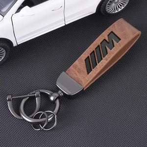 May include: A brown leather keyring featuring a black M logo embossed on the strap. The keyring includes a dark metallic hardware connector and a set of circular metal rings for holding keys, positioned next to a white toy car model.