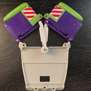 May include: A disassembled toy component, featuring a white base with a rectangular cutout and several circular holes. Attached are two purple and green wing-like structures with red and white striped accents. The toy is likely a part of a larger action figure.