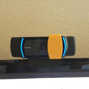 Puede incluir: Una webcam negra con detalles LED azules y un clip naranja. La webcam tiene un micrófono incorporado y está montada sobre una superficie negra. La webcam está etiquetada como "HD 1080p".