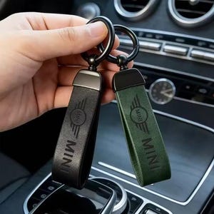 May include: Two leather keychains with black metal rings and hardware. One keychain is black and the other is dark green. Both feature the embossed MINI logo and the word MINI on the leather strap. The keychains are held together by a metal ring.