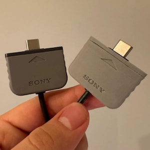 Könnte beinhalten: Zwei rechteckige Sony-Ladeadapter mit metallischen USB-C-Anschlüssen. Der linke Adapter ist dunkelgrau, der rechte hellgrau. Beide verfügen über ein eingeprägtes Dreieckssymbol über dem Sony-Logo und ein schwarzes Kabel, das nach unten führt.