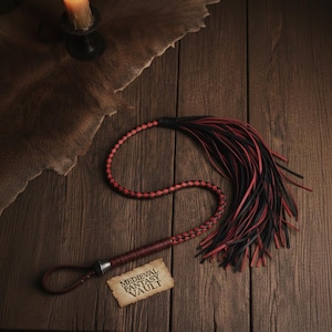 Puede incluir: Un látigo de cuero rojo y negro con un mango trenzado y un lazo en el extremo. El látigo tiene una serie de hebras de cuero negras y rojas en el extremo. Una pequeña etiqueta con el texto "MEDIEVAL FANTASY VAULT" está al lado del látigo.