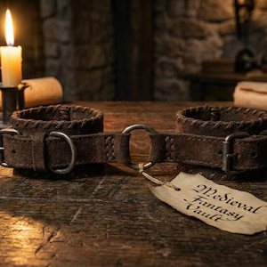 Puede incluir: Grilletes de cuero marrón con hebillas metálicas y un anillo de conexión, expuestos sobre una superficie de madera. Una vela encendida y un pergamino están en el fondo. Una etiqueta dice "Medieval Fantasy Vault".
