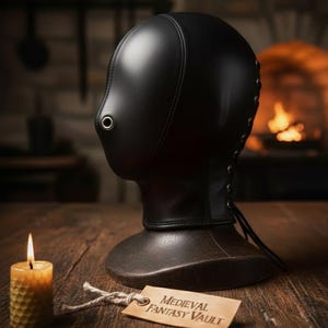 May include: A black leather hood with a metal eyelet and lace-up detailing. The hood sits on a dark brown base, with a lit candle and a tag that reads "Medieval Fantasy Vault". The background features a fireplace and wooden table.