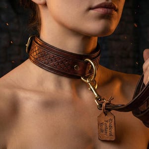 May include: A brown leather collar with a textured design and gold-colored hardware. A matching leash is held in the hand. A small tag with the text "Medieval Fantasy Vault" hangs from the collar.