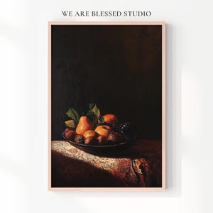 Op de afbeelding: Een ingelijste print met een stilleven van diverse vruchten in een donkere kom. De vruchten omvatten peren, sinaasappels en appels, met groene bladeren. De print is geplaatst tegen een donkere achtergrond en rust op een tafelkleed met patroon. De tekst "WE ARE BLESSED STUDIO" staat bovenaan.