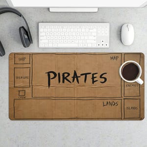 May include: A brown desk mat with a pirate map design. The mat features the word "PIRATES" in large black letters, along with labeled sections for "SHIP," "TREASURE," "CREW," "MAP," "ENEMIES," "LANDS," and "ISLANDS." A white coffee cup sits on the mat.