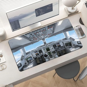 May include: A rectangular desk mat featuring a detailed image of an airplane cockpit interior, with various gauges, screens, and controls. The mat has a blue sky visible through the cockpit windows. The mat is on a white desk.