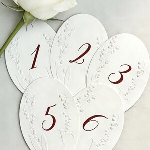 May include: White oval table number cards with embossed floral details and burgundy numbers 1 through 6. A white rose is visible in the upper left corner. Ideal for weddings or special events.