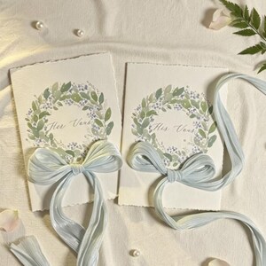 May include: Two invitation cards with watercolor floral wreaths and the text "Her Verse" and "His Verse". Each card is adorned with a light blue ribbon bow. The cards are on a white surface with scattered pearls, rose petals, and greenery.