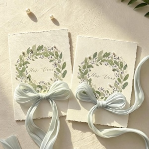 May include: Two wedding cards with watercolor floral wreaths and the text "Her Vows". Each card is adorned with a light blue ribbon bow. Scattered pearls and greenery add to the elegant, romantic aesthetic.