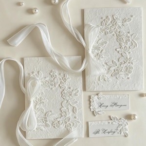 May include: Two rectangular white textured paper cards featuring white floral lace appliqués, small pearl accents, and long sheer white ribbons tied in bows. Two smaller rectangular name cards with black cursive text and lace details are included.