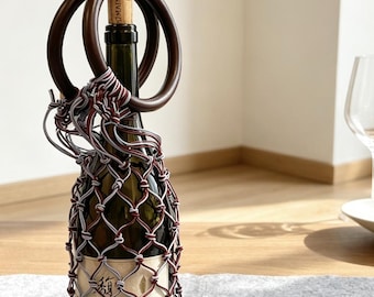 May include: A wine bottle encased in a decorative macrame net carrier featuring a diamond-shaped pattern made of maroon and light gray cord. The carrier includes two circular dark brown wooden handles at the top, secured around the bottle neck.