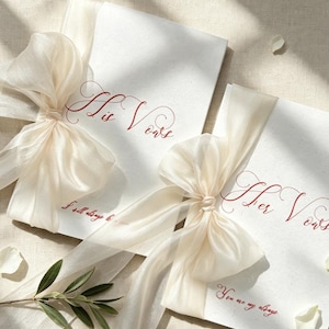 May include: Two white rectangular vow booklets featuring red cursive text. One booklet reads His Vows and I will always be yours, the other reads Her Vows and You are my always. Each booklet is tied with a sheer cream-colored ribbon bow.