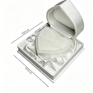 Pu&ograve; includere: Un portagioie bianco a forma di cuore con interno foderato in raso ed esterno a coste. La scatola presenta dimensioni di 90 mm per 97 mm, con un inserto interno a forma di cuore di 65 mm per 70 mm, indicate da linee di misurazione nere.