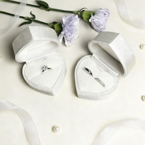 Pu&ograve; includere: Due scatole per anelli a forma di cuore bianco con coperchi testurizzati, una contenente un anello solitario con diamante e l'altra due fedine in argento. Le scatole sono disposte con due garofani color viola chiaro e nastri bianchi.