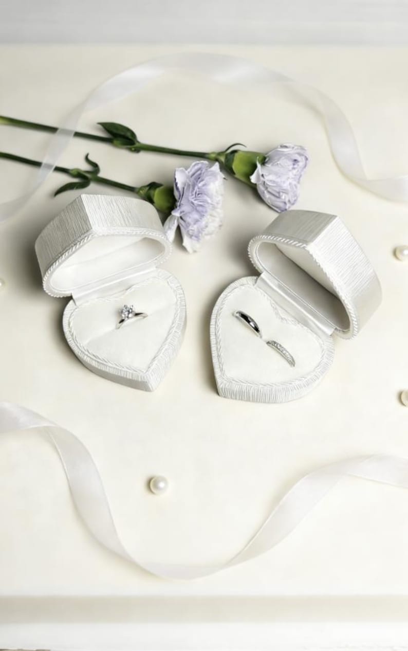 Pu&ograve; includere: Due scatole per anelli a forma di cuore bianco con coperchi testurizzati, una contenente un anello solitario con diamante e l'altra due fedine in argento. Le scatole sono disposte con due garofani color viola chiaro e nastri bianchi.