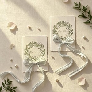 May include: Two wedding vow cards with a green wreath design and the text "Her Vows". Each card has a light blue ribbon tied in a bow. Scattered around are pearls, rose petals, and greenery.