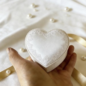 Pu&ograve; includere: Un portagioie a forma di cuore con la parte superiore in raso bianco decorata da un delicato bordo in pizzo bianco e una fascia laterale a coste bianche. La scatola &egrave; compatta e presenta un profilo dai bordi lisci e arrotondati.