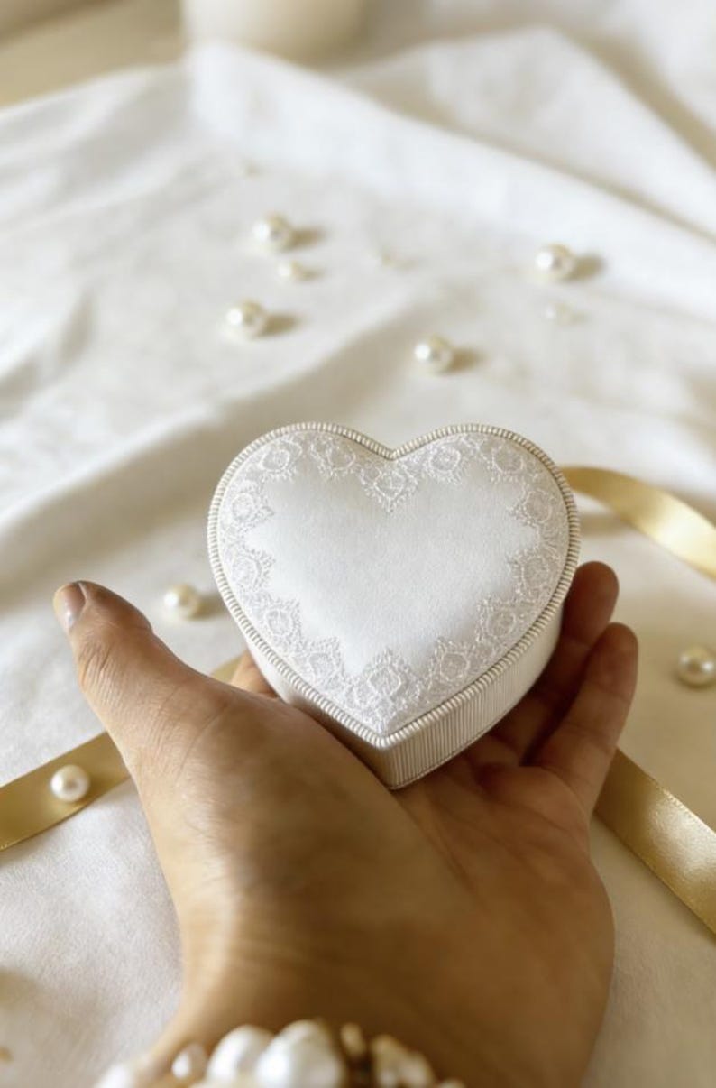 Pu&ograve; includere: Un portagioie a forma di cuore con la parte superiore in raso bianco decorata da un delicato bordo in pizzo bianco e una fascia laterale a coste bianche. La scatola &egrave; compatta e presenta un profilo dai bordi lisci e arrotondati.