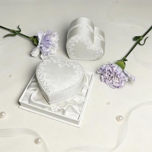 Pu&ograve; includere: Due scatole per anelli bianche a forma di cuore con lati a coste e motivi floreali in pizzo sui coperchi. Una scatola poggia all'interno di una base quadrata bianca foderata in raso, mentre l'altra si trova accanto, accompagnata da due garofani color malva.