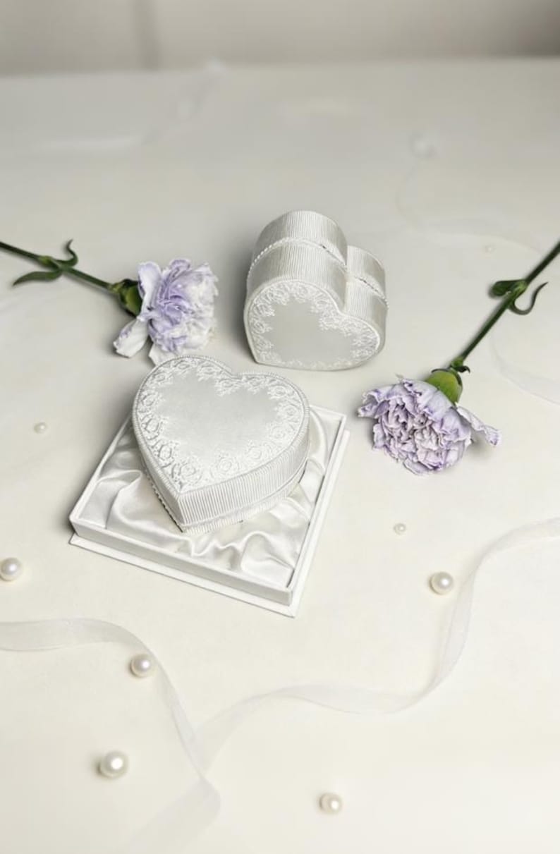 Pu&ograve; includere: Due scatole per anelli bianche a forma di cuore con lati a coste e motivi floreali in pizzo sui coperchi. Una scatola poggia all'interno di una base quadrata bianca foderata in raso, mentre l'altra si trova accanto, accompagnata da due garofani color malva.
