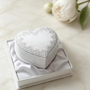 Pu&ograve; includere: Un portagioie bianco a forma di cuore con un lato a coste e un coperchio decorato con un intricato ricamo in pizzo bianco. La scatola &egrave; inserita all'interno di un contenitore di presentazione quadrato bianco foderato in morbido raso bianco.