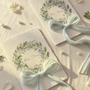May include: Two ivory-colored cards with watercolor floral wreath designs and the text "Her Vows". Each card is adorned with a light blue ribbon bow and scattered pearls, creating an elegant, romantic aesthetic.