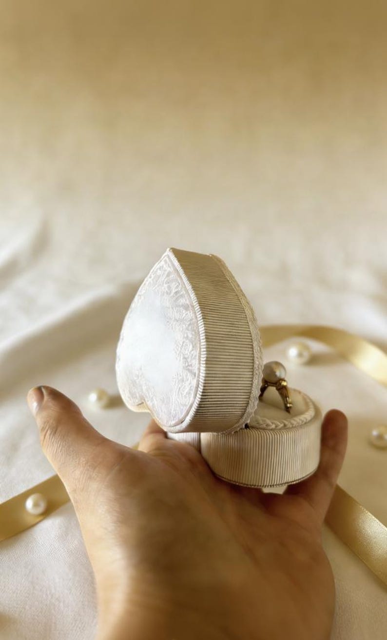 Pu&ograve; includere: Un portagioie a forma di cuore con esterno a coste color crema e coperchio con motivo in pizzo bianco. All'interno della scatola aperta si trova un anello d'oro con una singola perla bianca. La scatola &egrave; decorata con un nastro color crema.