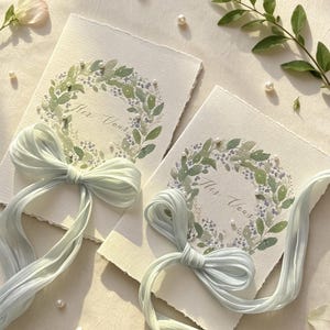 May include: Two invitation cards with watercolor floral wreaths and the text "Her Vous". Each card is adorned with a light blue ribbon bow. Scattered pearls and greenery add to the elegant design.