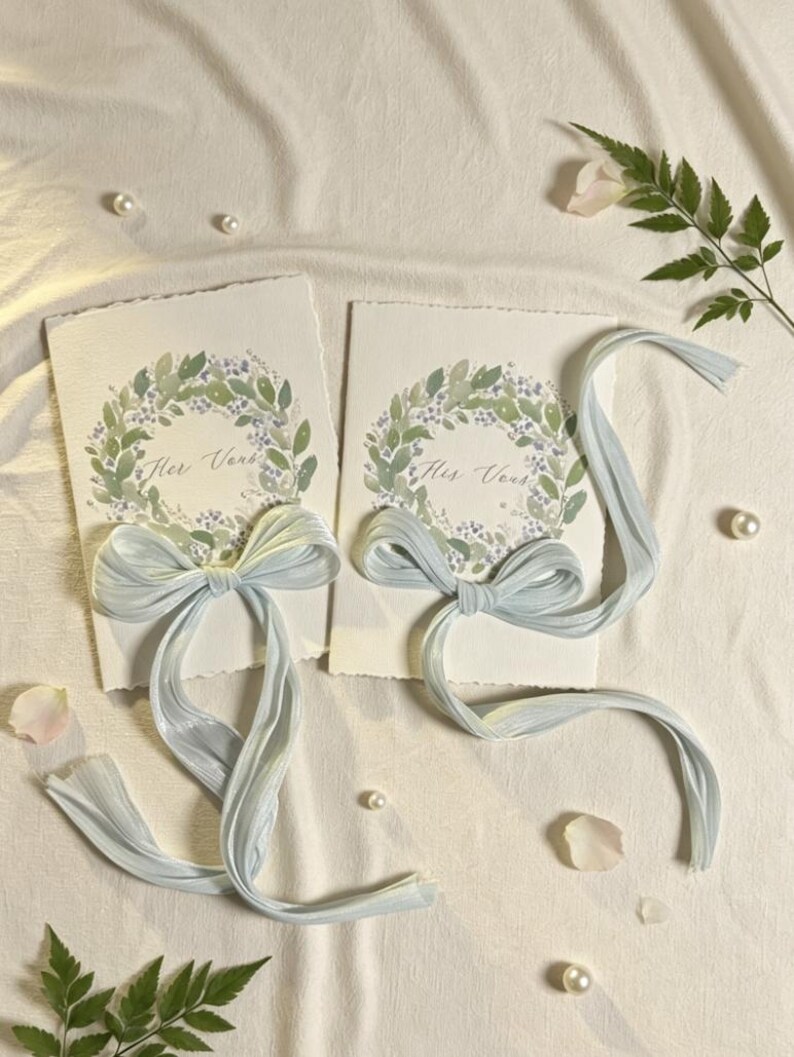 May include: Two invitation cards with watercolor floral wreaths and the text "Her Verse" and "His Verse". Each card is adorned with a light blue ribbon bow. The cards are on a white surface with scattered pearls, rose petals, and greenery.