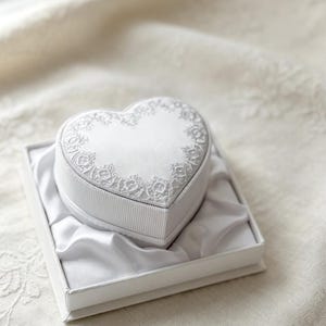 Pu&ograve; includere: Un portagioie bianco a forma di cuore con la parte superiore in raso decorata da un intricato ricamo in pizzo bianco lungo i bordi. I lati della scatola presentano una trama a coste ed &egrave; inserita in una scatola quadrata foderata in raso.