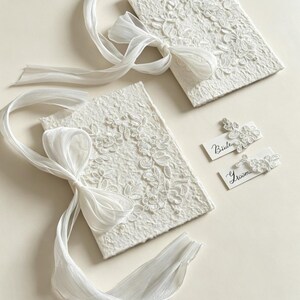 May include: Two rectangular white textured paper booklets adorned with white floral lace appliques and long sheer ribbons tied in bows. Two small white rectangular cards are included, one labeled Bride and one labeled Groom, each featuring a matching lace accent.