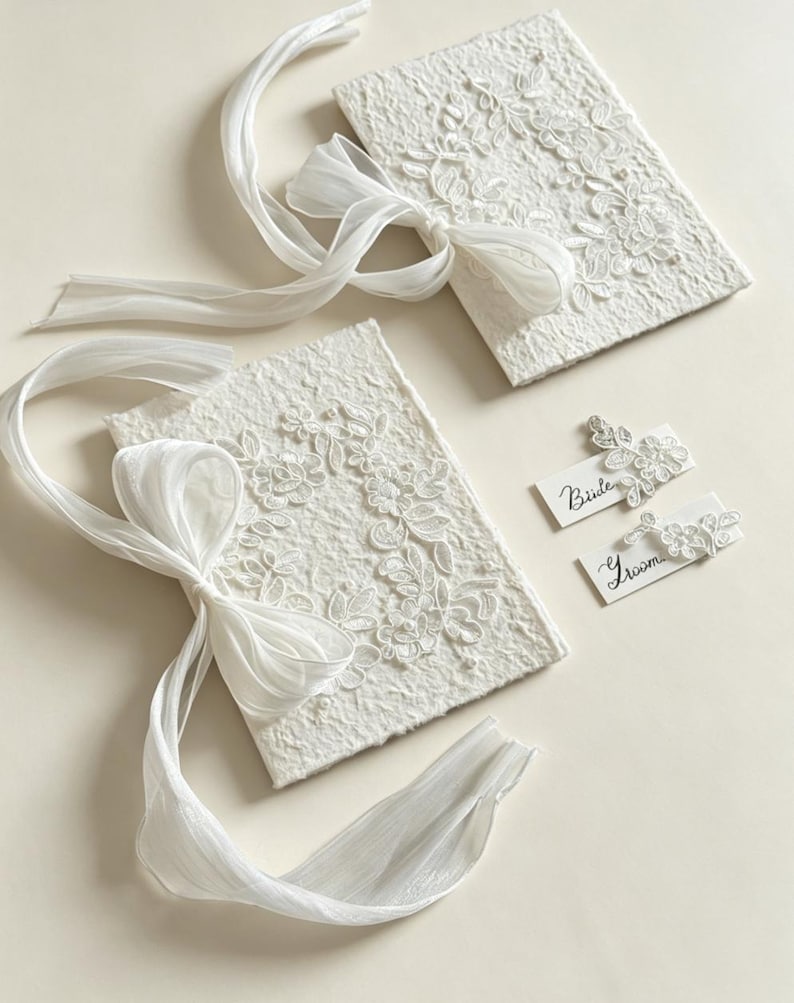May include: Two rectangular white textured paper booklets adorned with white floral lace appliques and long sheer ribbons tied in bows. Two small white rectangular cards are included, one labeled Bride and one labeled Groom, each featuring a matching lace accent.