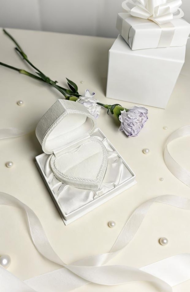 Pu&ograve; includere: Un portagioie bianco a forma di cuore con coperchio testurizzato e interno foderato in raso. Il portagioie &egrave; aperto e rivela un inserto in velluto a forma di cuore per un anello. &Egrave; presentato con una scatola regalo bianca e un nastro.