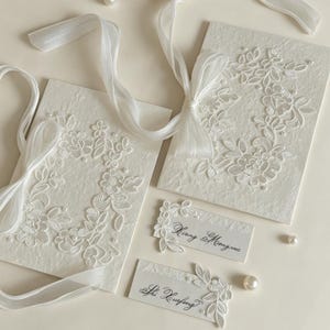 May include: Two off-white textured paper cards featuring white floral lace appliqu&eacute;s, sheer white ribbon bows, and small pearl accents. Two smaller rectangular cards are included, one with the text Xiang & Mengxue and one with Shi Xuefeng.